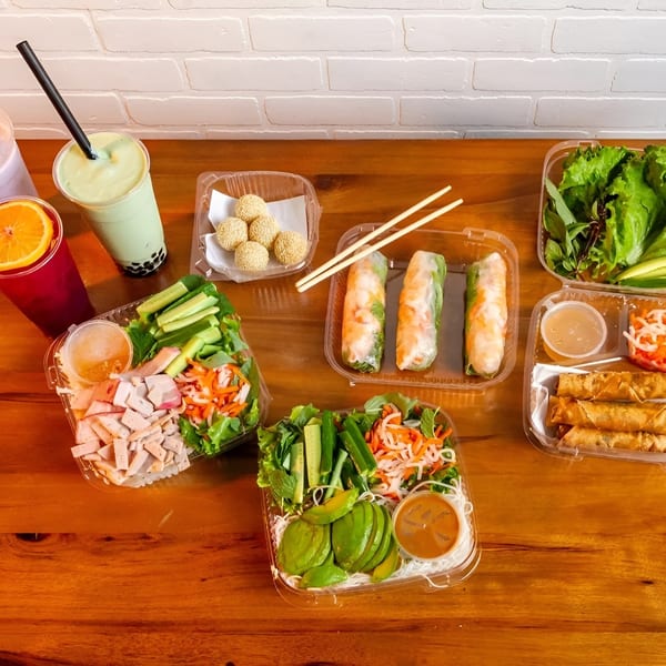 Assortment of Vietnamese dishes in takeout containers, including spring rolls, salads, and drinks.